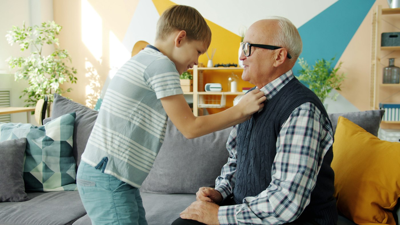 Helpful grandson helping elderly grandfather with clothing while old man is having fun playing with kid at home. Caring people, lifestyle and family concept.